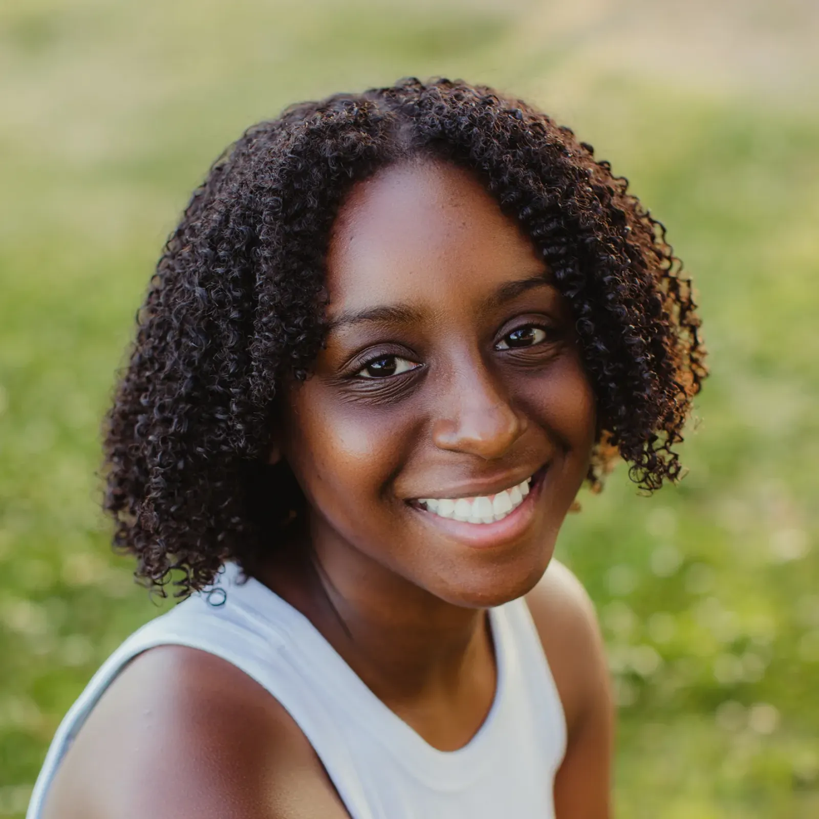 Smiling Black woman with curly hair and a white sleeveless top poses outdoors, warmly lit against a soft, green blurred background.
