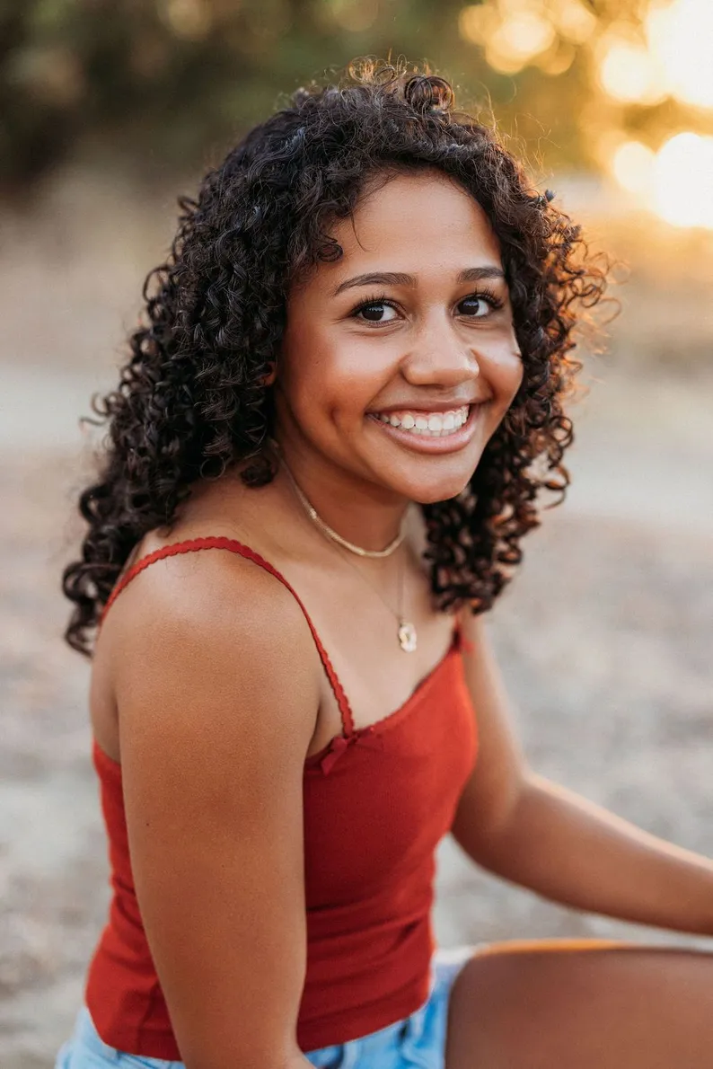 Smiling woman with curly hair in a red top sits outdoors at golden hour, her joyful expression framed by warm, softly blurred sunlight.