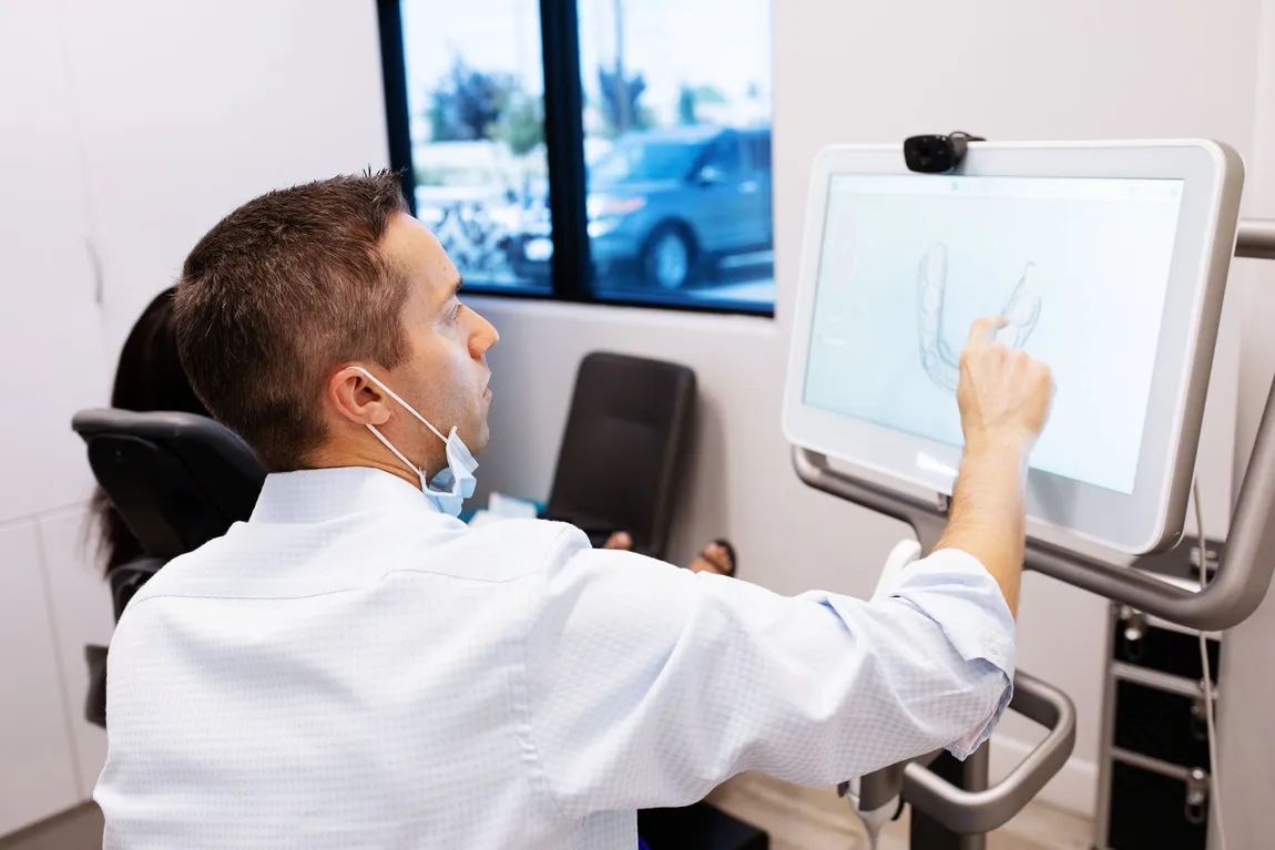 A healthcare professional in a white coat and face mask uses a touchscreen medical monitor in a bright clinic, suggesting diagnosis or care.