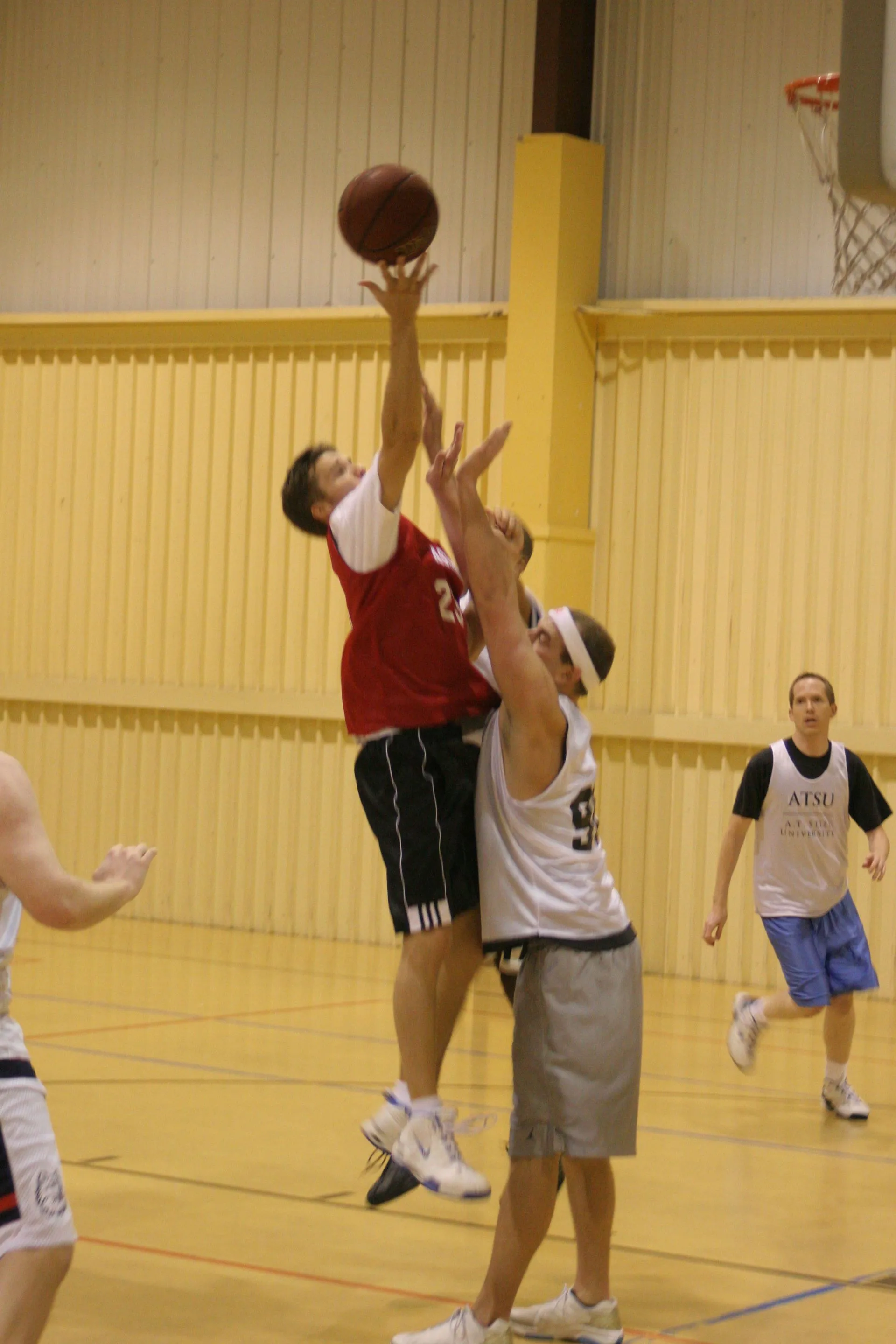 Basketball players compete indoors as a red-uniformed player reaches for the ball near the hoop, challenged by a defender; teammates watch.