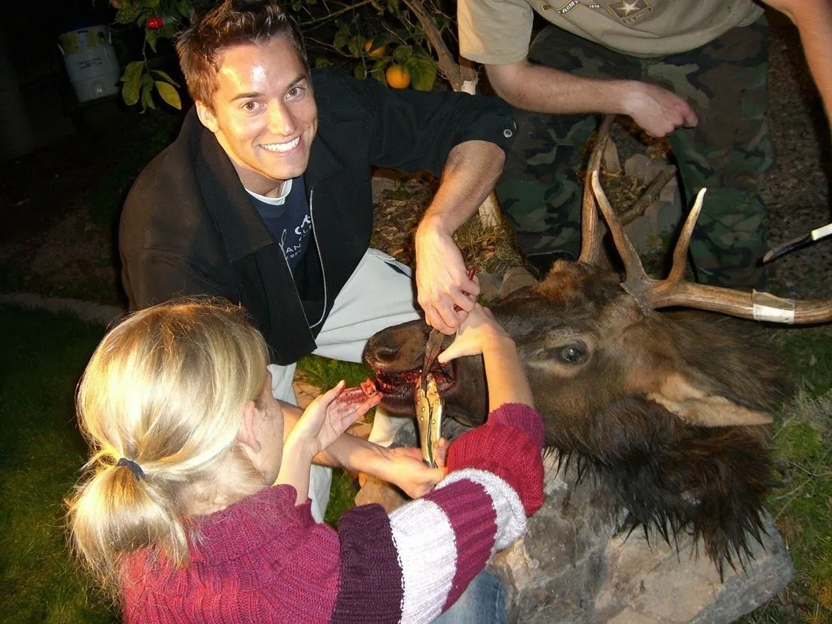 Smiling man and woman examine a large antlered deer trophy outdoors at night, creating a lively hunting-themed scene.
