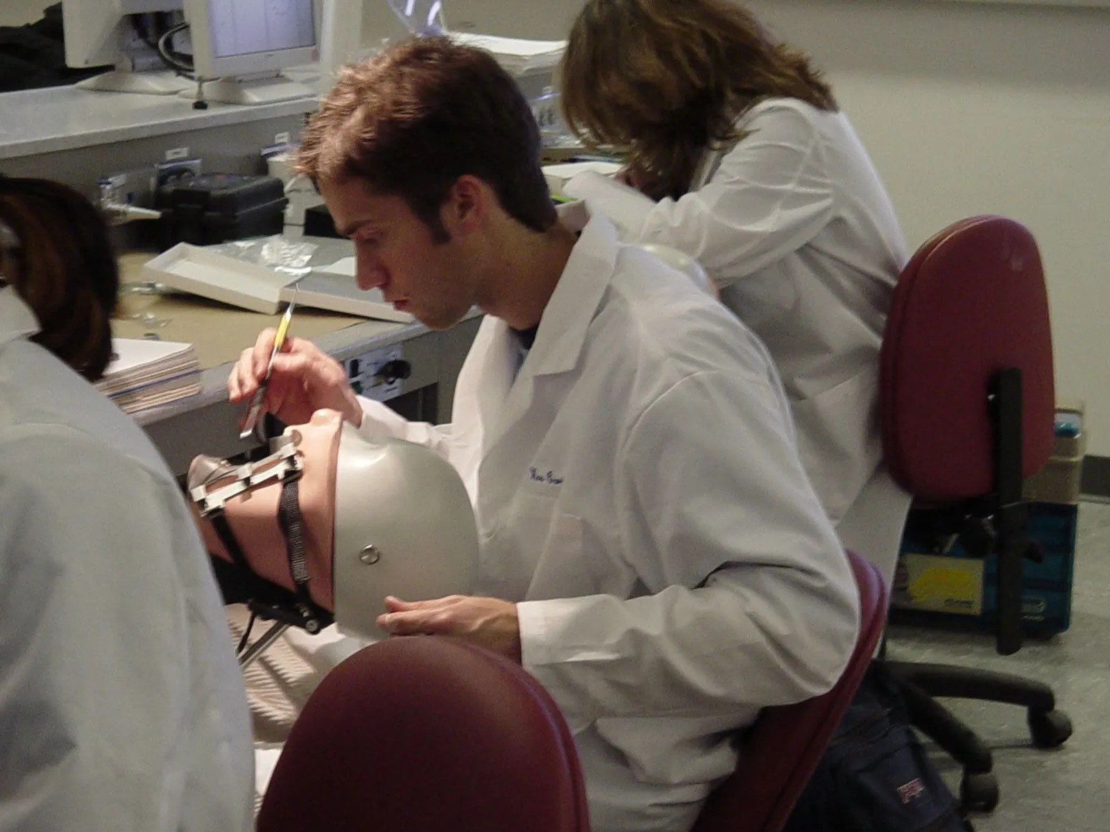 Dental students in white lab coats practice dentistry on a mannequin head in a classroom lab, creating a focused, hands-on learning scene.