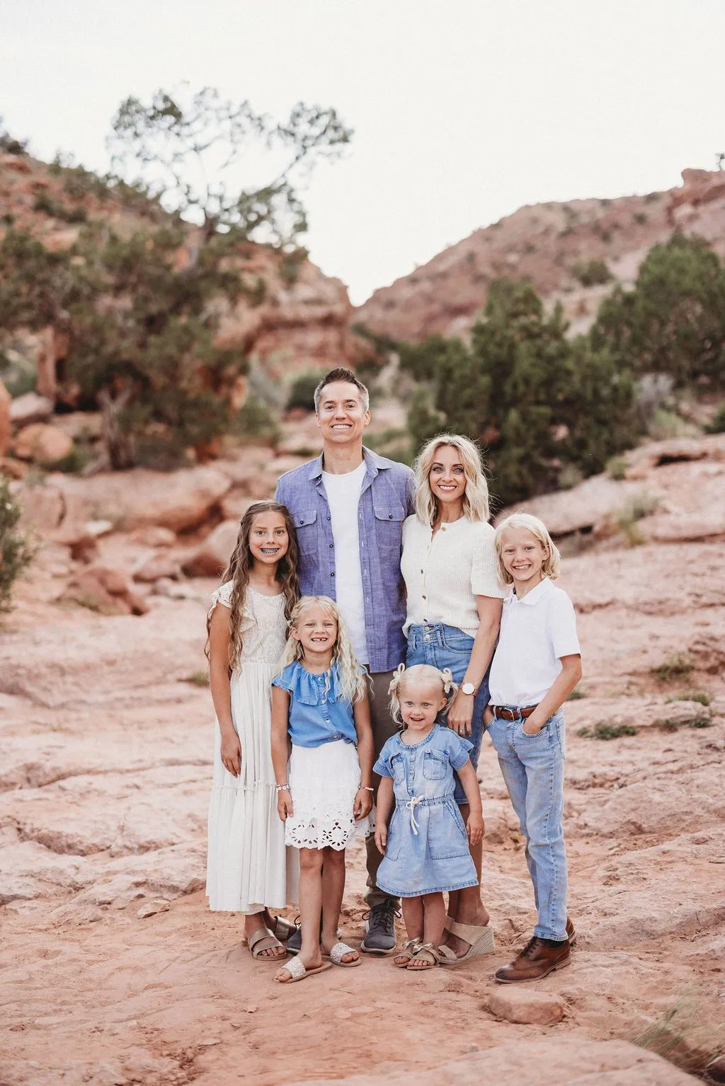 Smiling family of six poses together on a rocky desert trail, surrounded by red canyon walls and trees in warm, peaceful light.