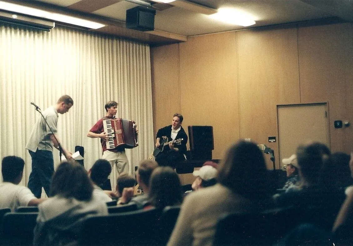 Three musicians perform an intimate acoustic concert—accordion, guitar, and percussion—for a seated audience in a warmly lit room.
