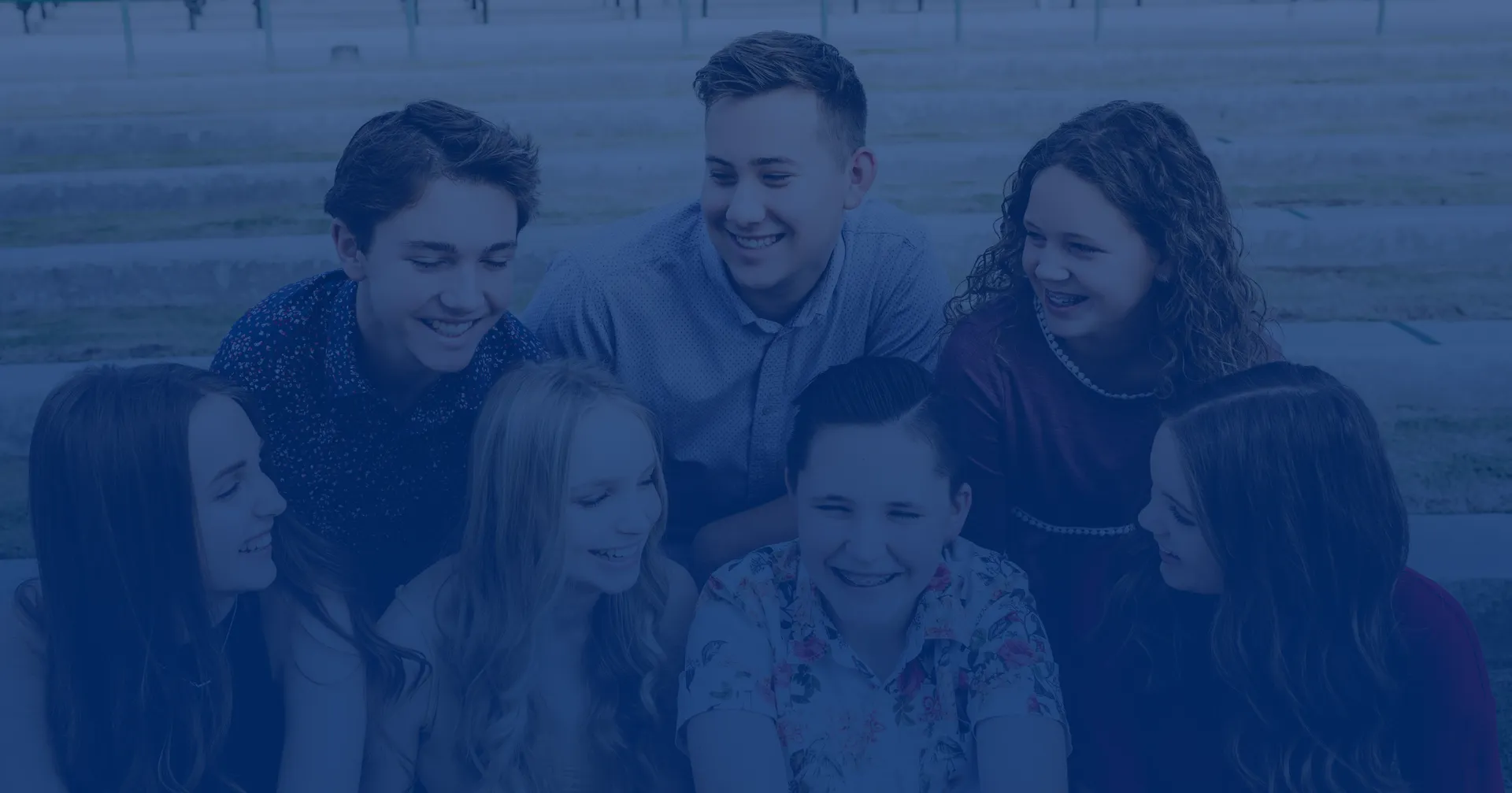 Seven young friends gather outdoors, smiling and laughing together in a close group portrait, conveying warmth, friendship, and joyful connection.