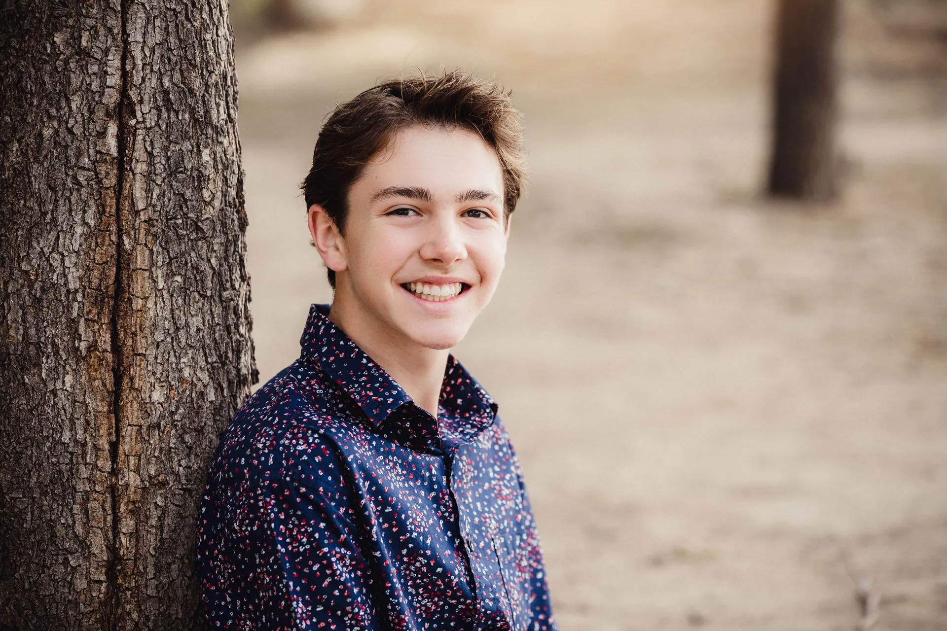 Smiling teenage boy in a colorful patterned shirt leans beside a tree in a sunlit park, conveying warmth and cheerful confidence.