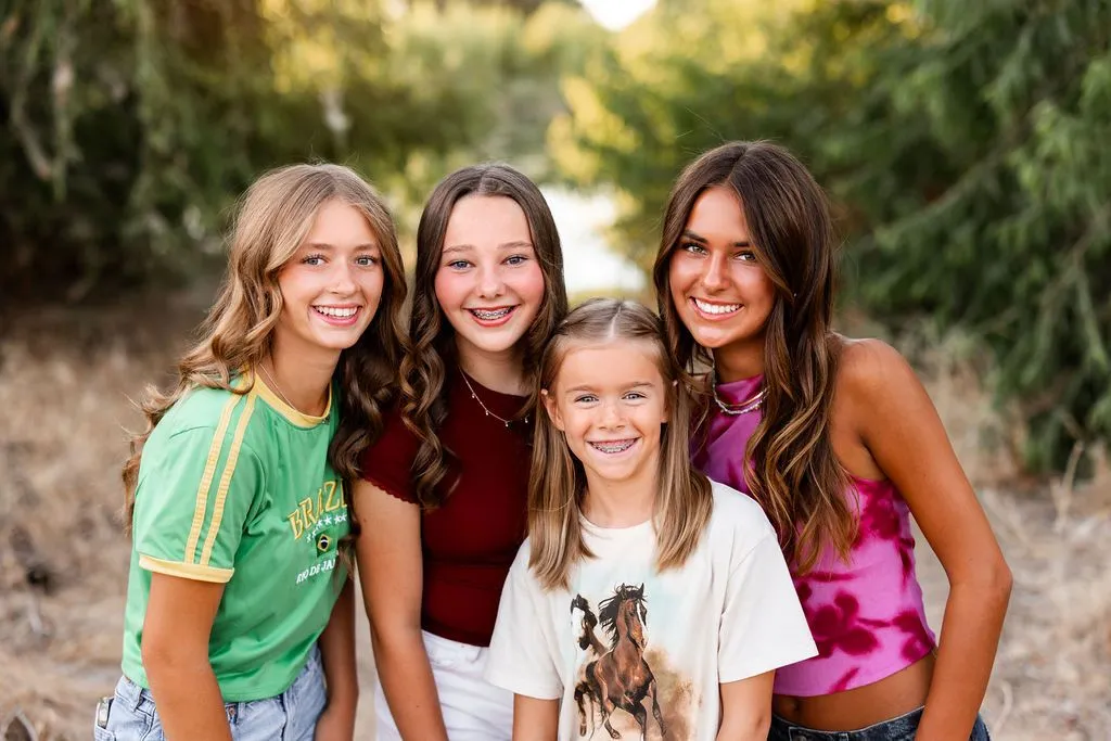 Four smiling girls pose together outdoors on a wooded path, wearing colorful casual clothes and showing a cheerful, friendly mood.