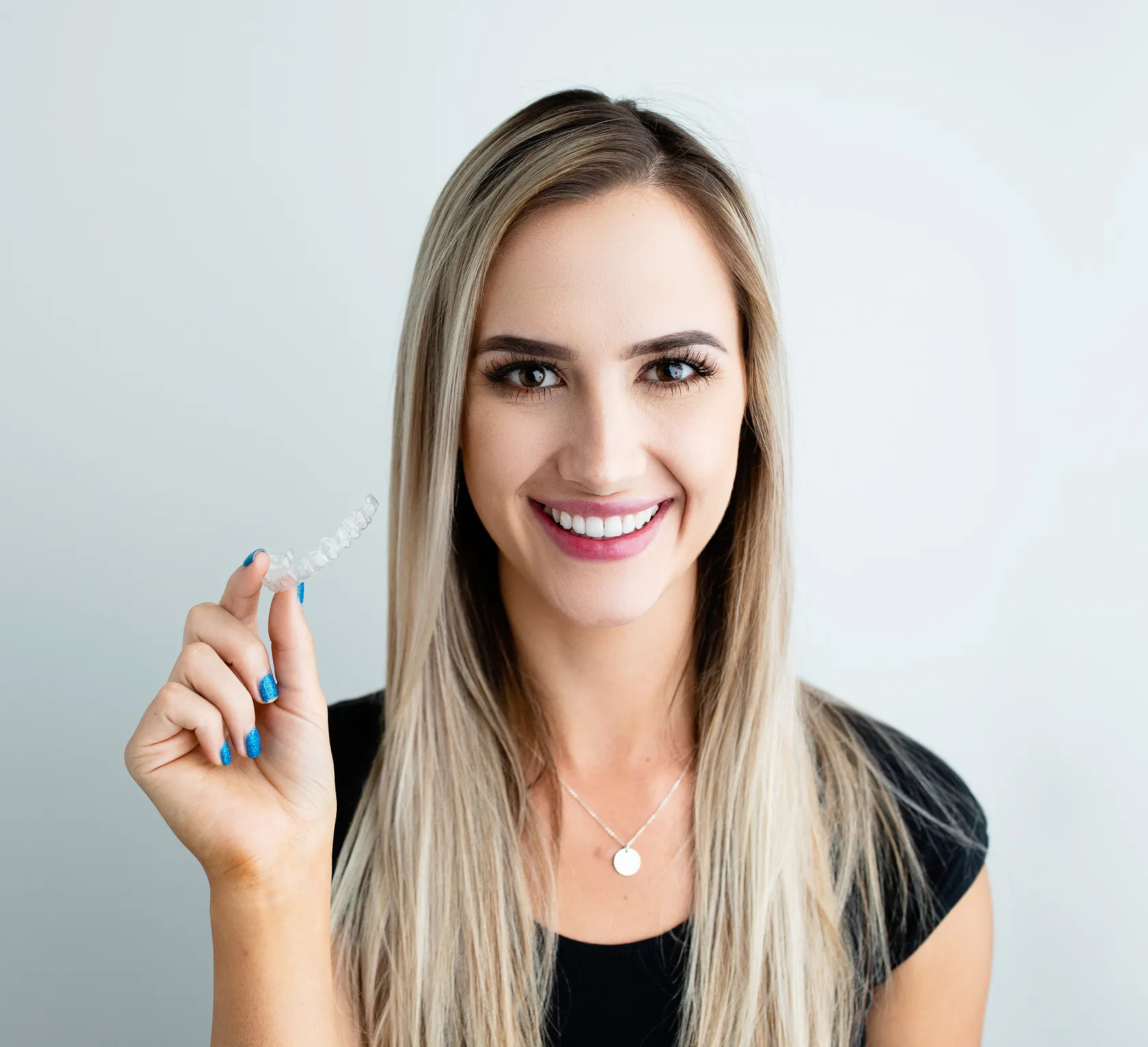 Smiling blonde woman in a black top holds a clear dental aligner beside her face, showcasing a bright, confident orthodontic smile.