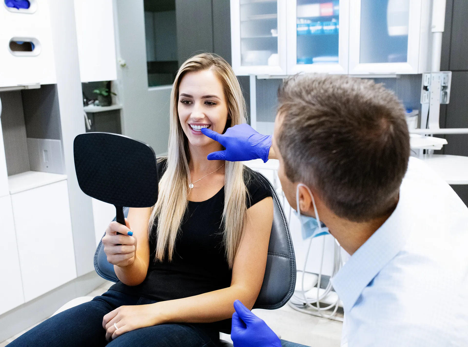 Dentist in blue gloves examines a smiling woman’s teeth as she holds a mirror in a modern dental clinic, conveying friendly, reassuring care.