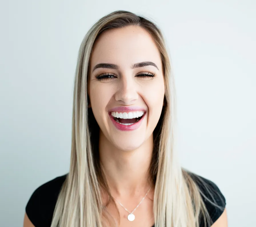 Portrait of a blonde woman in a black top laughing brightly against a pale background, eyes nearly closed and white teeth visible; joyful mood.