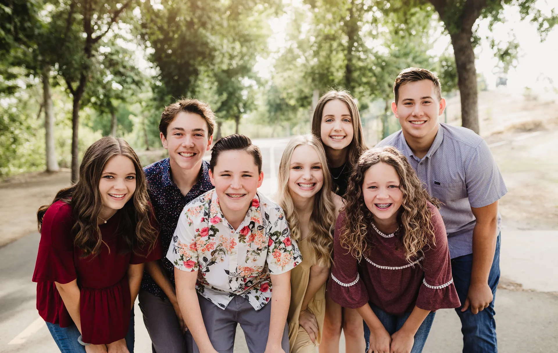 Seven smiling friends pose together outdoors in a sunlit park, leaning toward the camera in a cheerful, relaxed, warm group portrait.