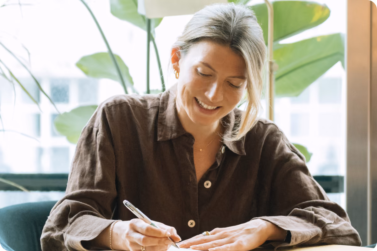 Woman in a brown linen shirt smiling while signing a document at a desk, with green plants in the background
