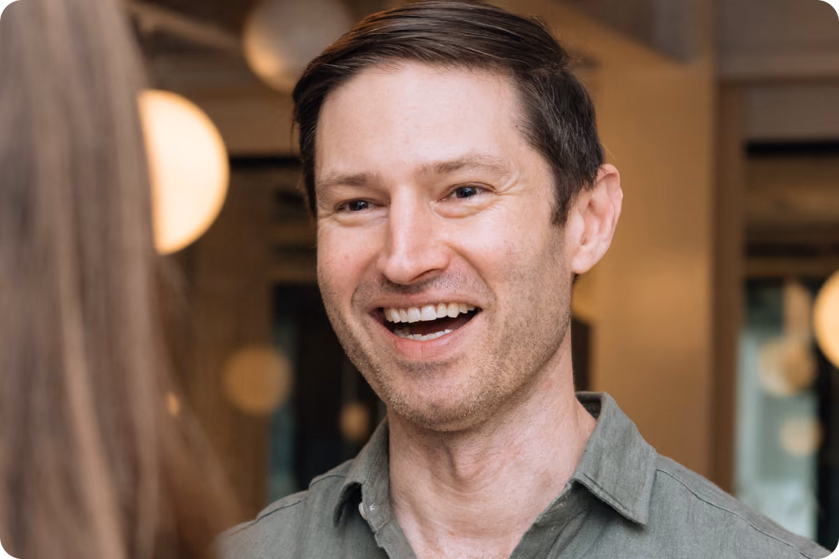 Man in an olive shirt laughing during a conversation with a colleague, office with string lights in the background