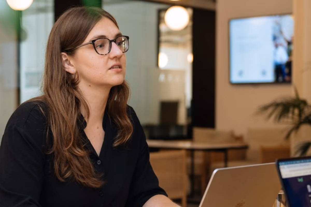 Woman with glasses working on a laptop at a wooden table in a modern office, with a second laptop screen visible in the foreground