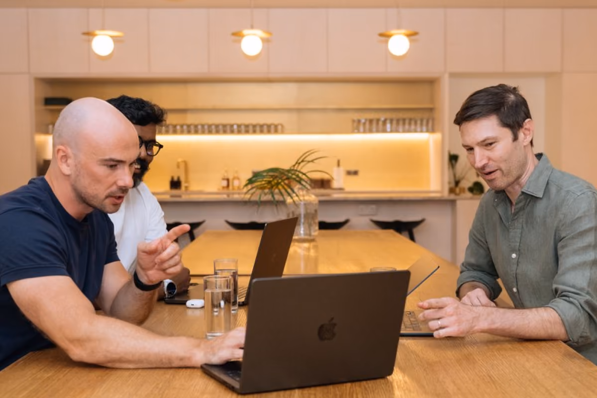 Three colleagues gathered around laptops in conversation at a wooden table in an office kitchen area