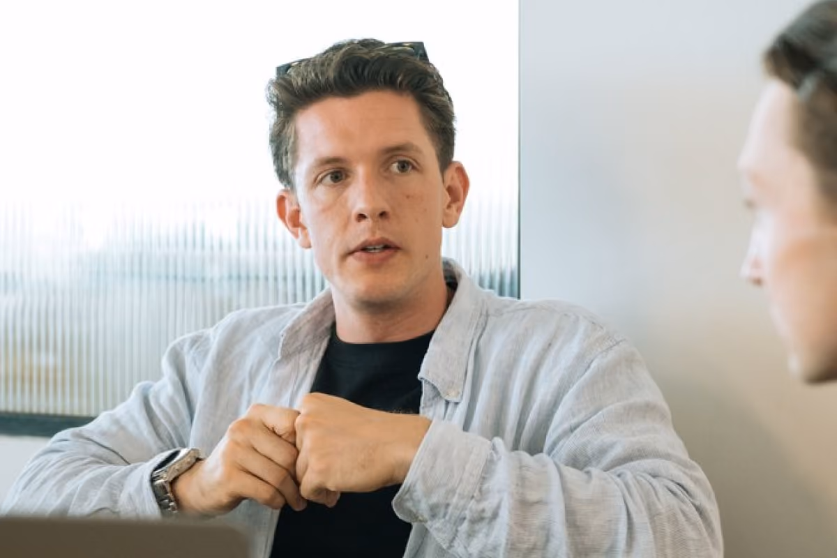 Man in a light jacket gesturing while talking with a colleague, laptop open on the table in front of them