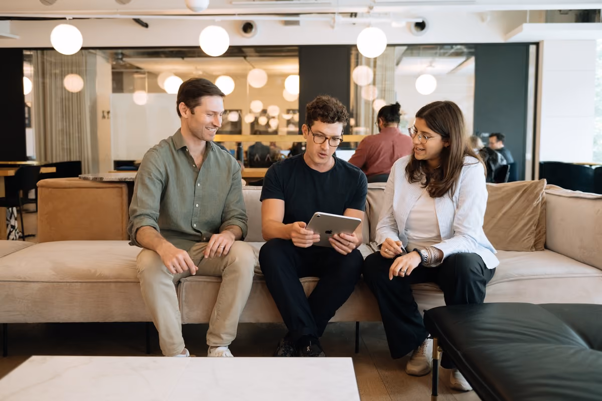 Three colleagues sitting on a sofa reviewing content on a tablet in a modern office space.