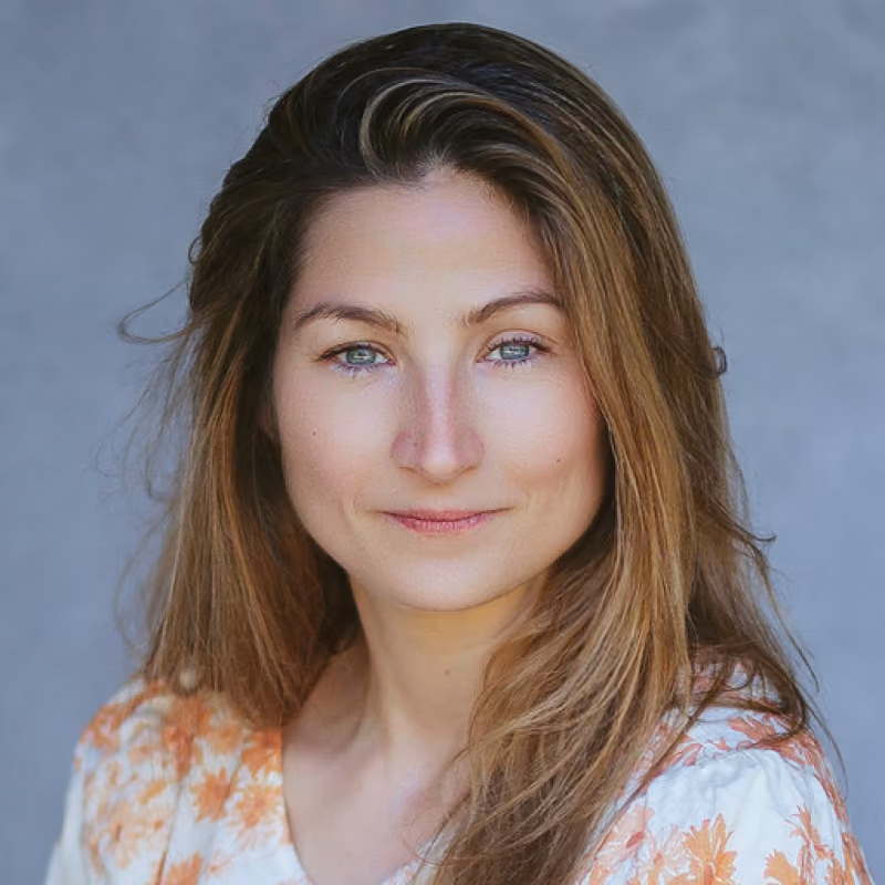 Young woman with long light brown hair in a floral blouse smiling gently, grey background.