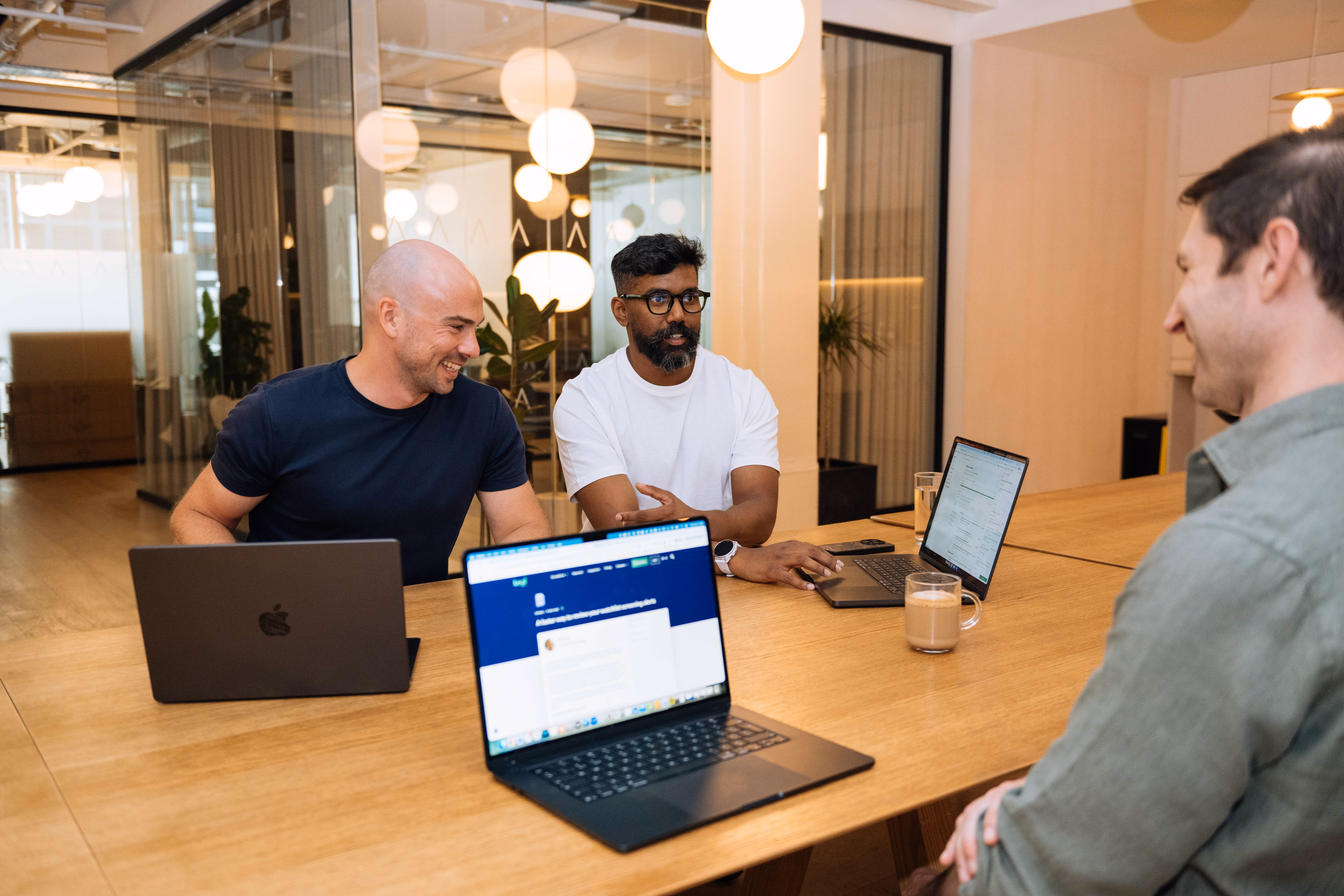 Three men having a meeting with laptops at a large wooden table in a modern office space.