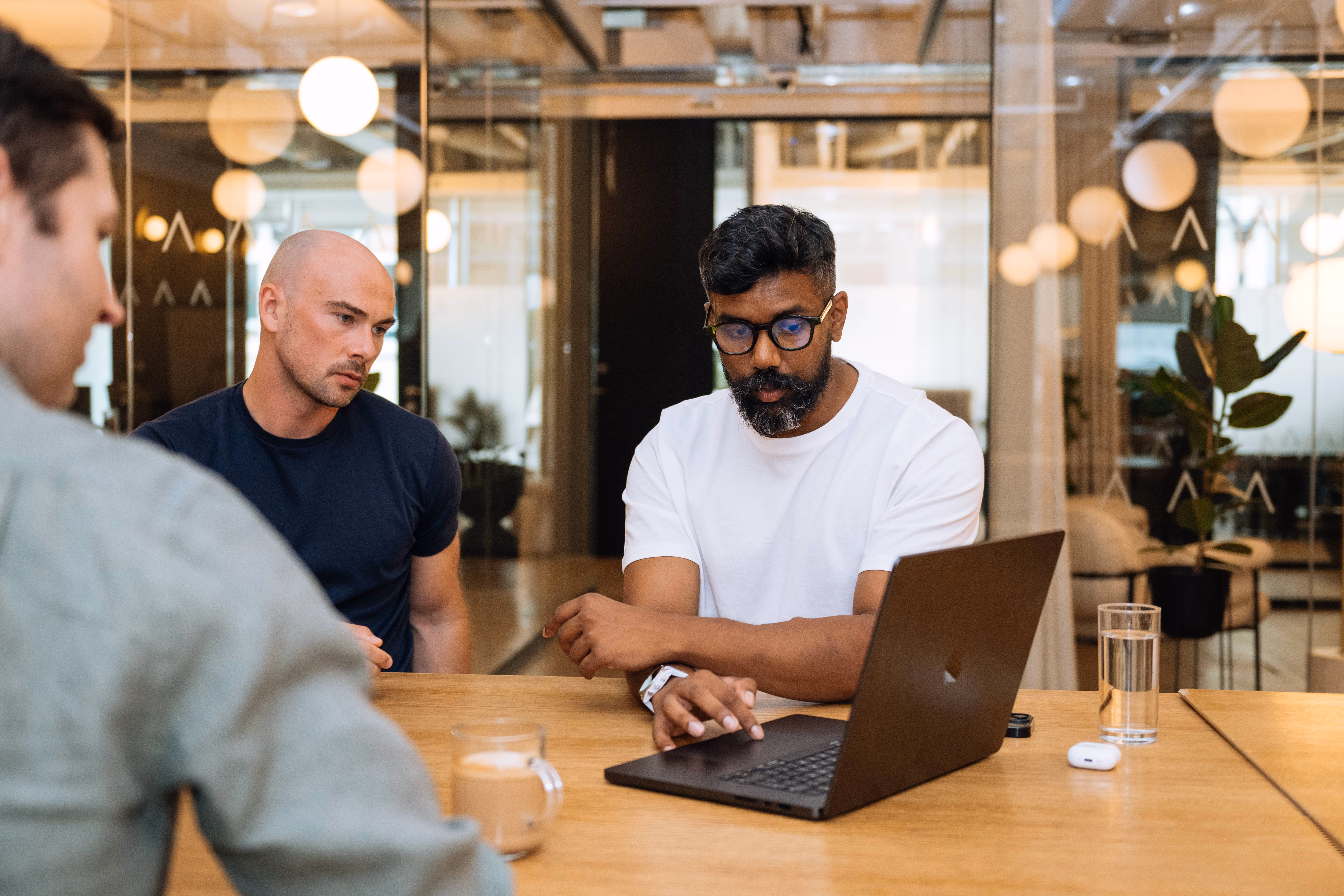 Three men in a meeting room focused on a laptop during a discussion at a wooden table.