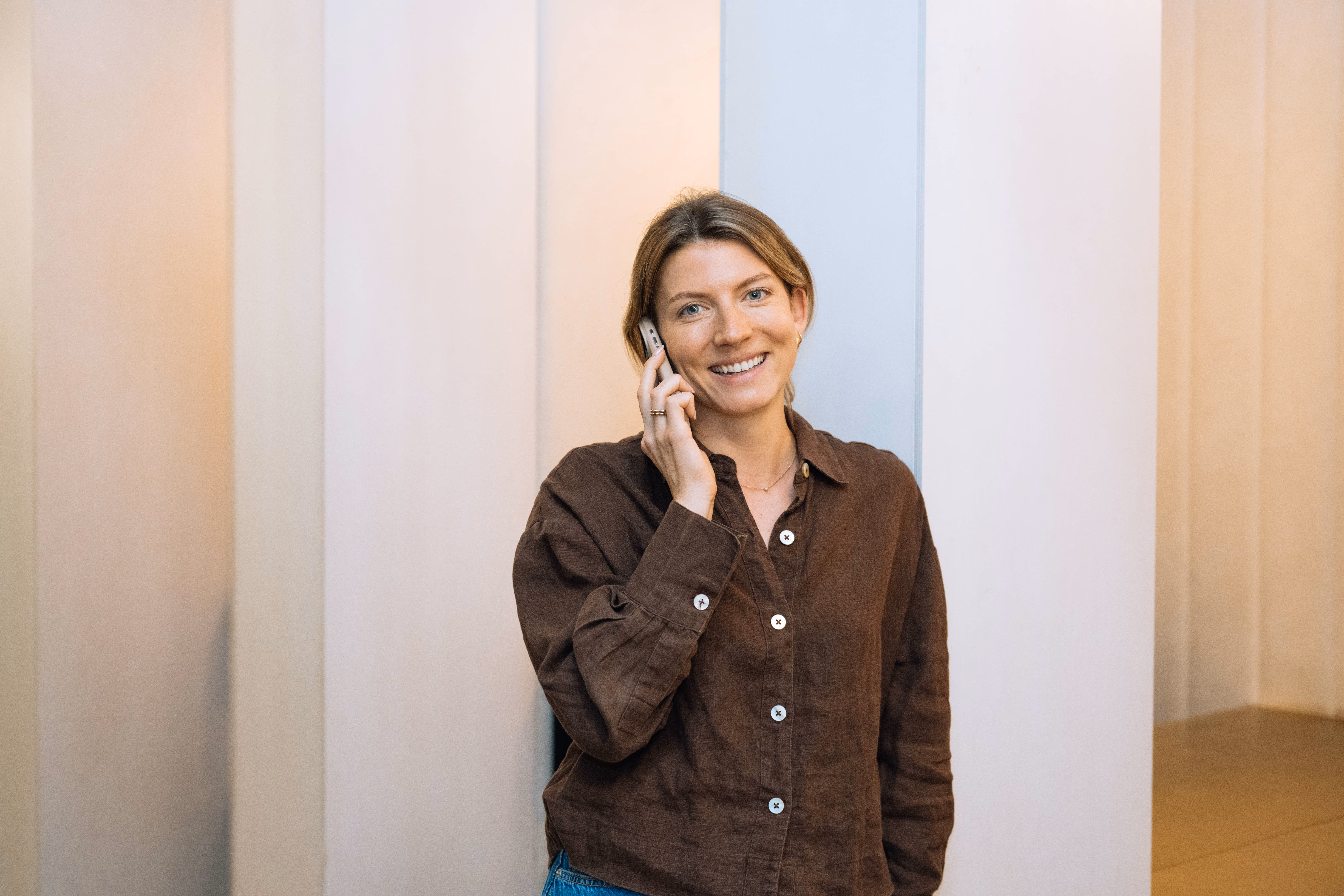 Smiling woman with brown shirt talking on a mobile phone against a light grey and beige wall.