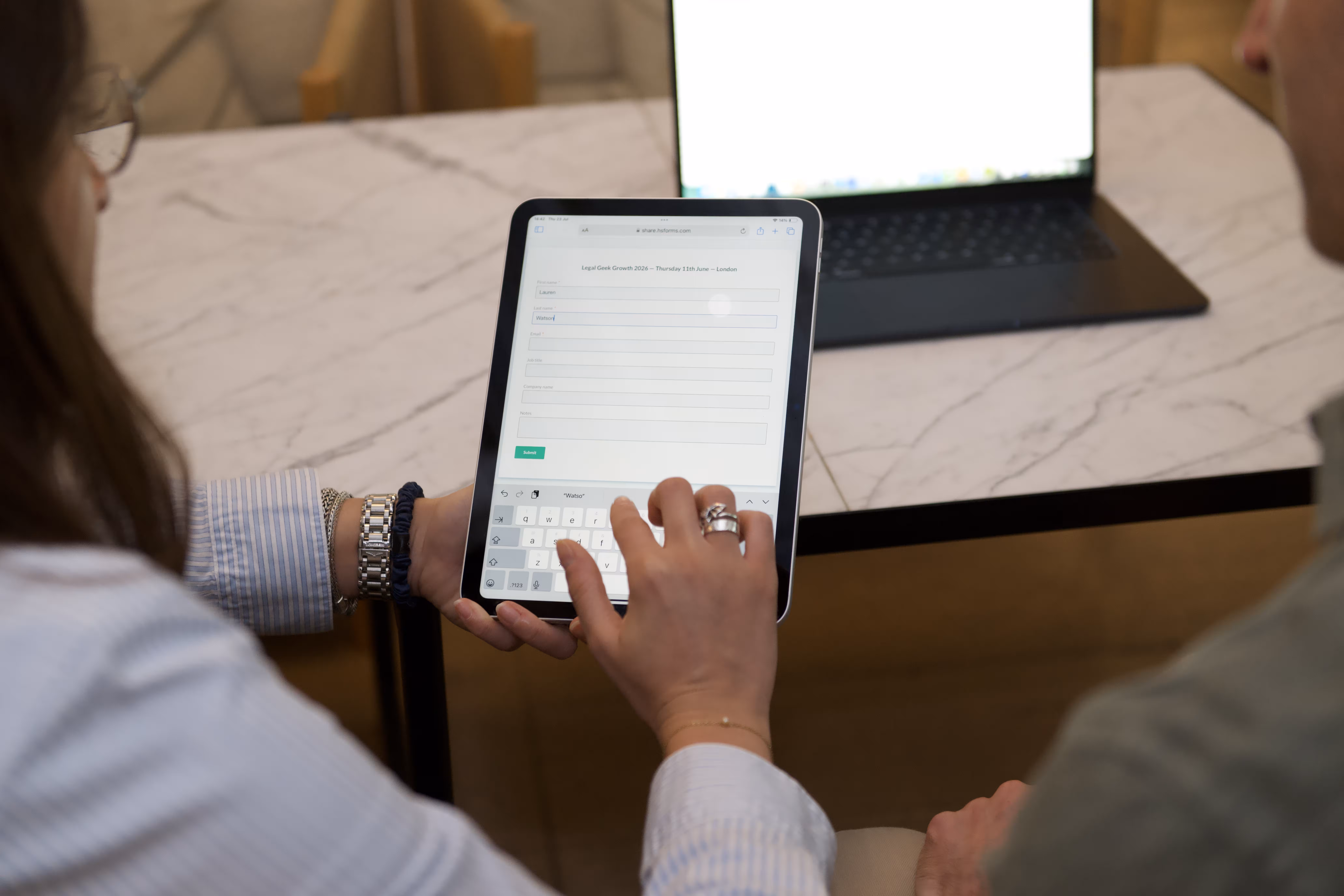 Person typing on a tablet filling a legal event form while another looks on, with a laptop on a marble table.