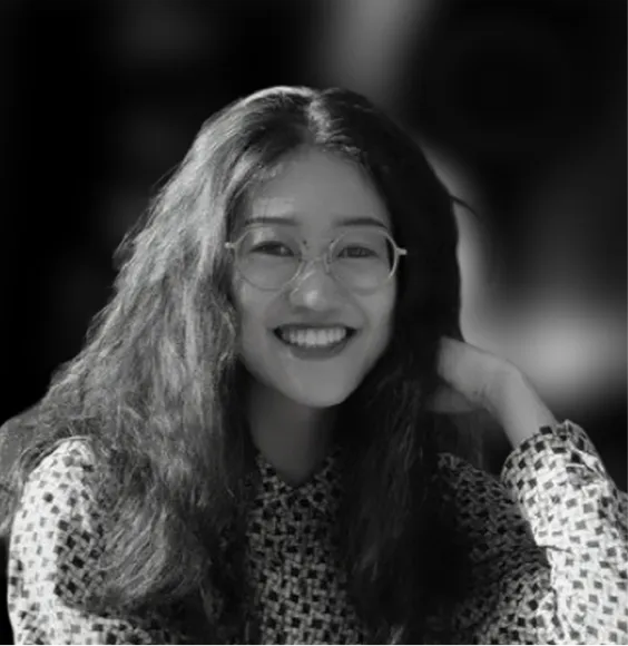 Smiling woman with long hair and glasses wearing a patterned top with hand near her head.