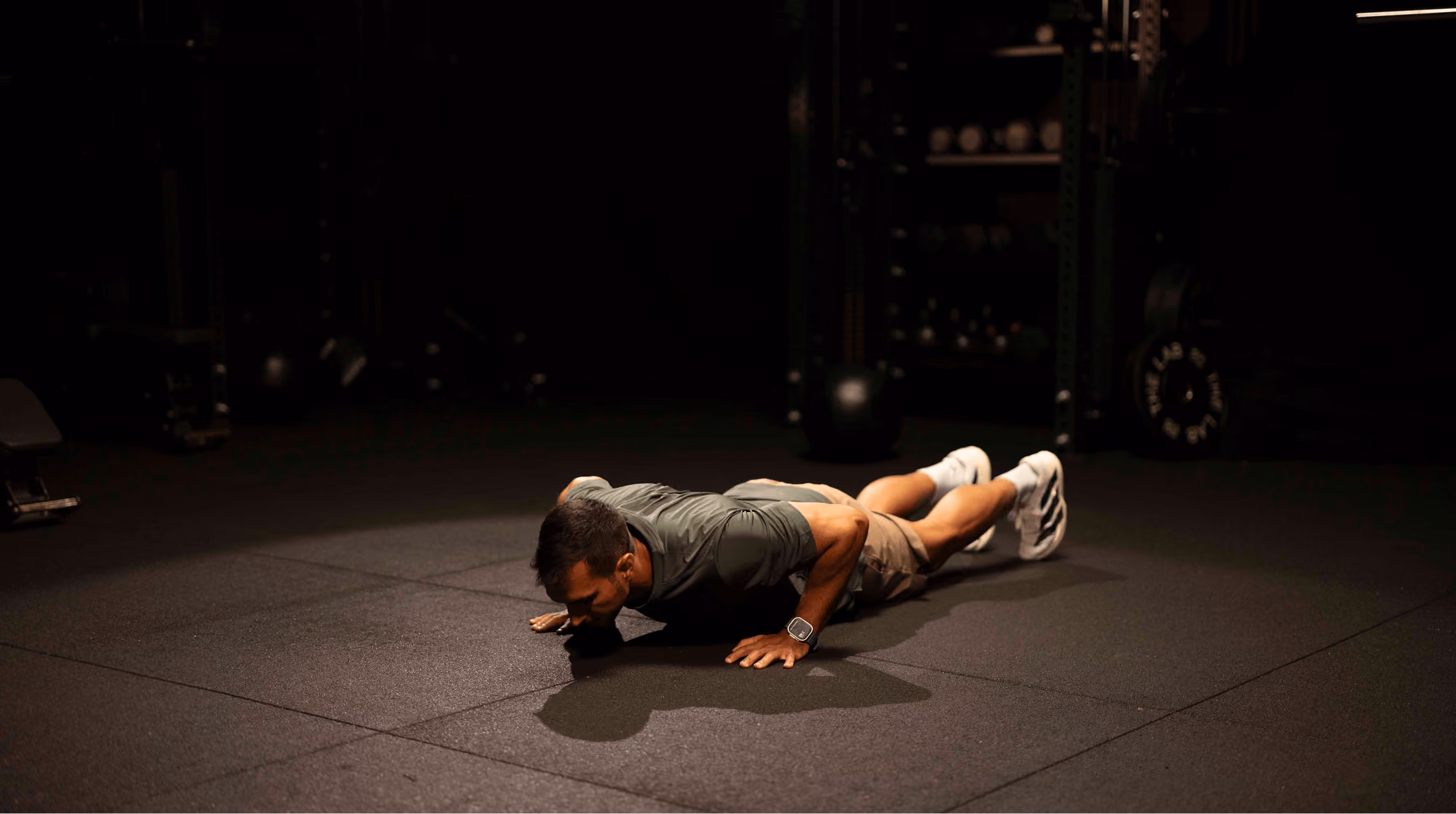 Man in athletic wear doing a push-up on a gym floor with dark gym equipment in the background.