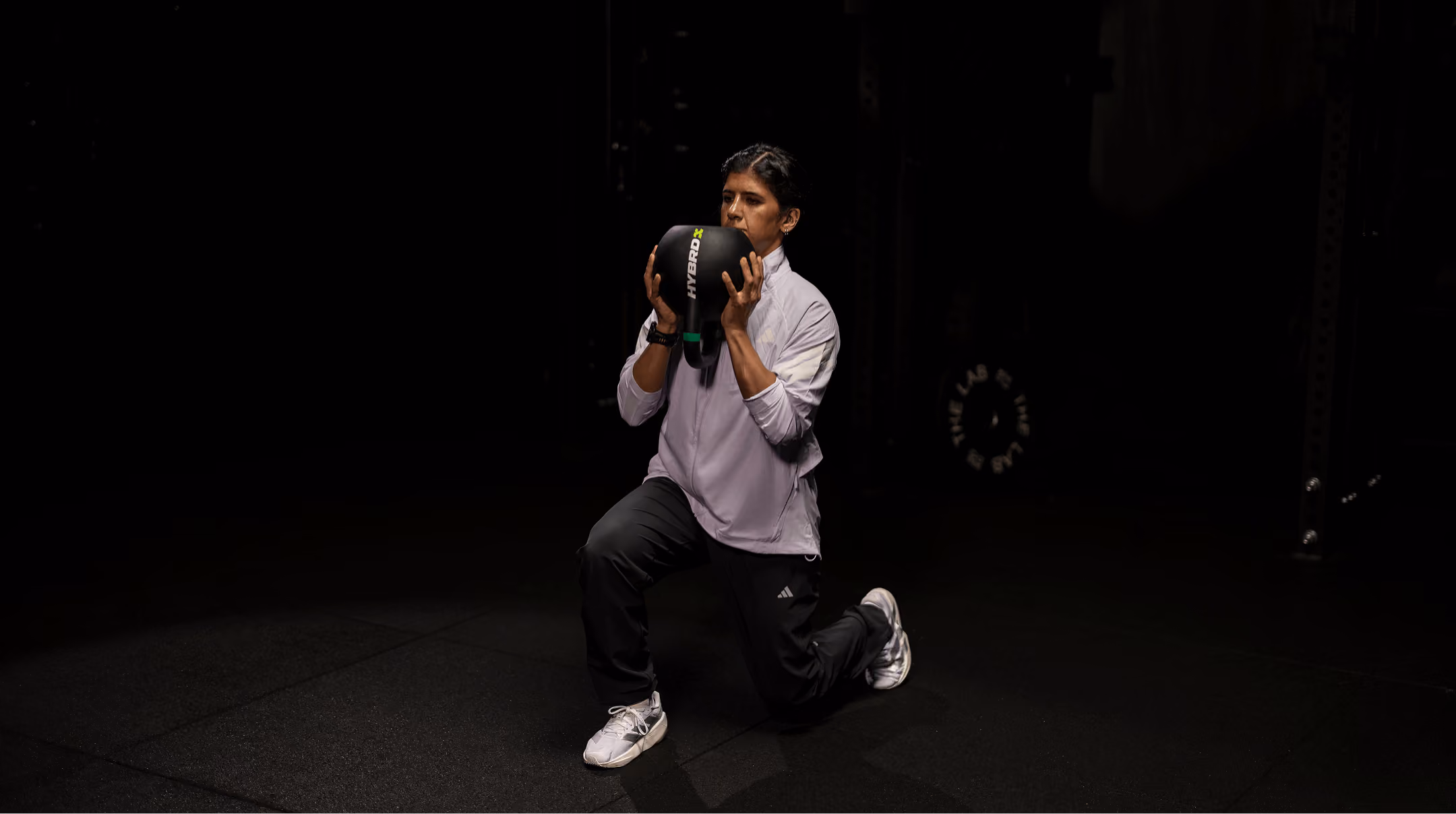 Person in a light purple jacket and black pants doing a lunge while holding a black kettlebell labeled HYBRD.