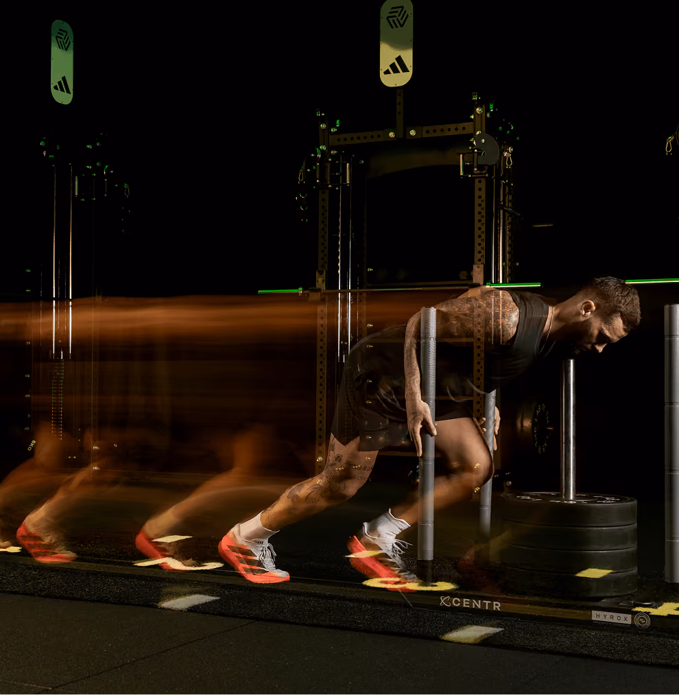 Athletic man with tattoos wearing black shorts and a sleeveless top pushes a weighted sled forward in a gym, wearing bright red sneakers, captured with motion blur showing his movement.