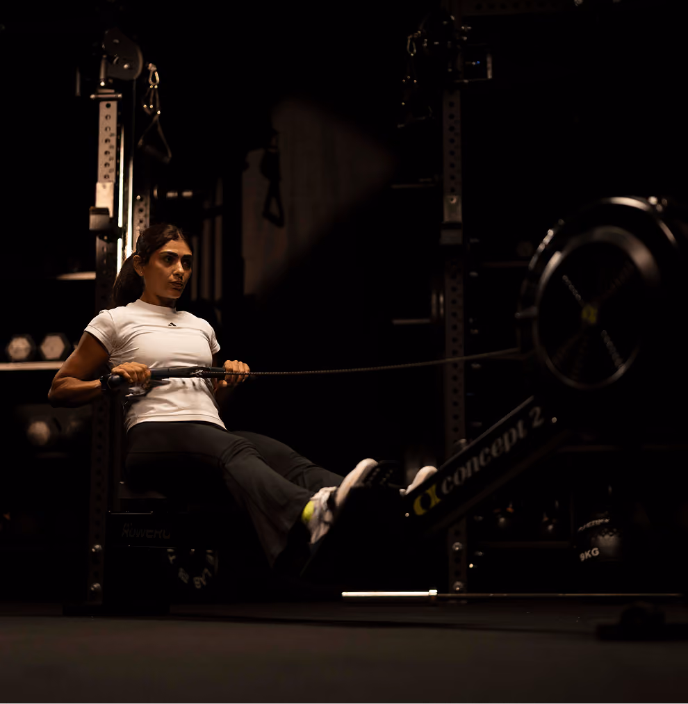 Woman wearing a white shirt and black workout pants using a Concept2 rowing machine in a dimly lit gym.