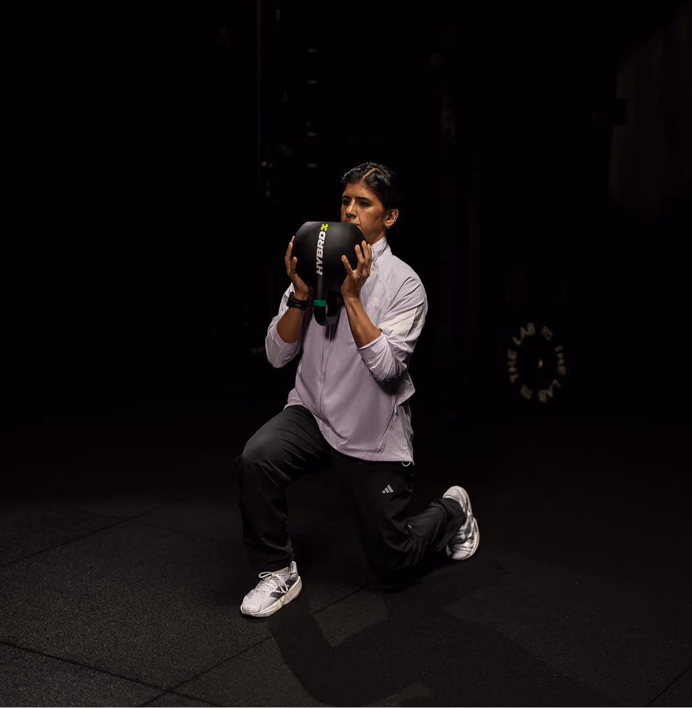 A woman in a light jacket and dark pants performs a kneeling lunge while holding a black HYBRD kettlebell close to her chest in a dimly lit gym.