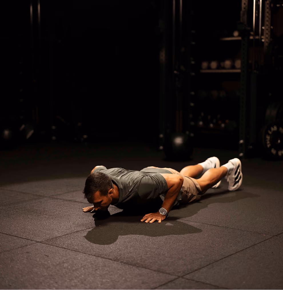 Man wearing a dark green shirt, tan shorts, and white athletic shoes performing a close-to-ground push-up on a black gym floor in a dimly lit fitness facility.