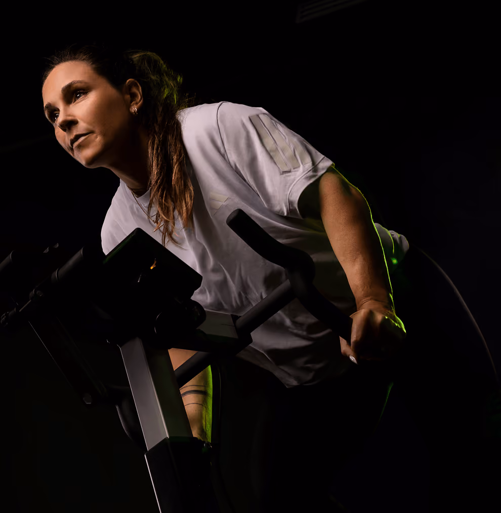 Focused woman in a white shirt leaning forward while using a stationary exercise bike in a dark setting with green accent lighting.