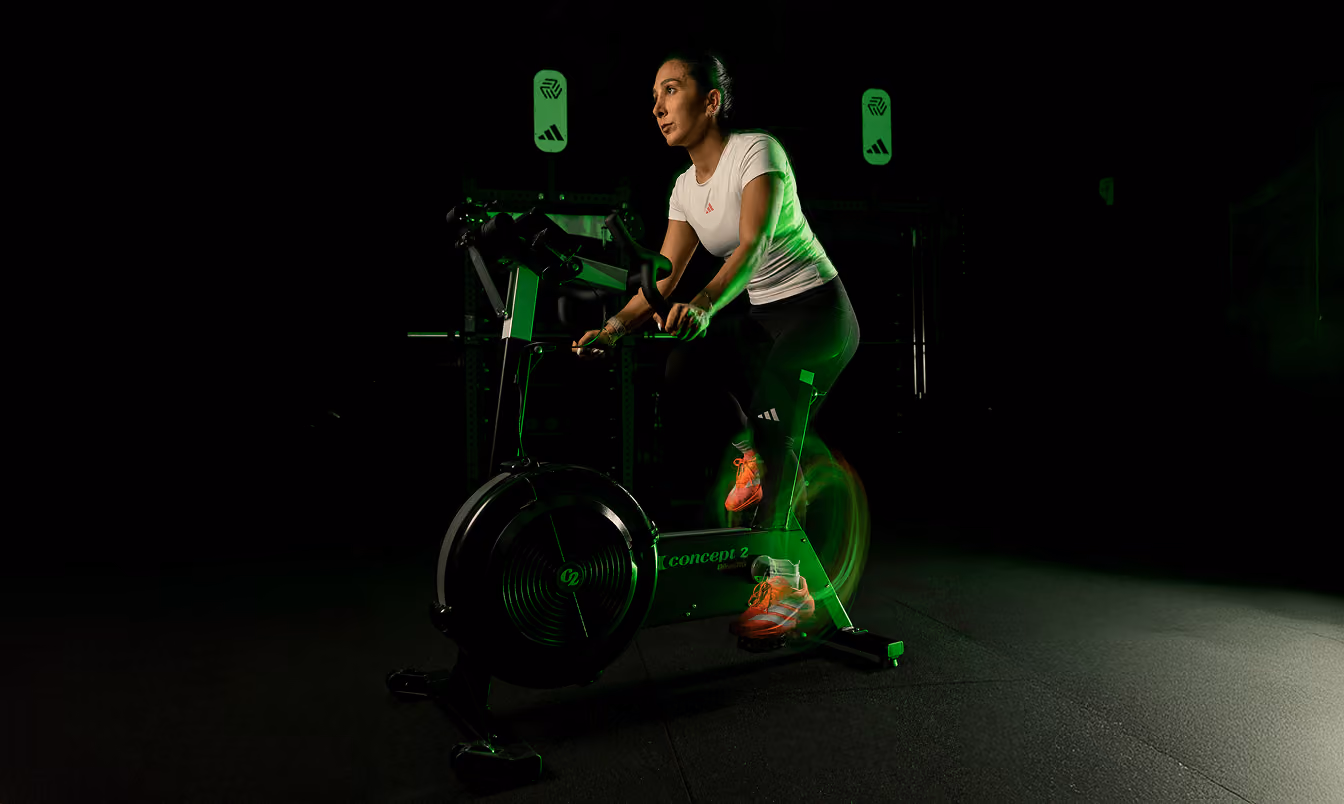 Woman in a white shirt and black leggings riding a stationary exercise bike in a dark room with green lighting.
