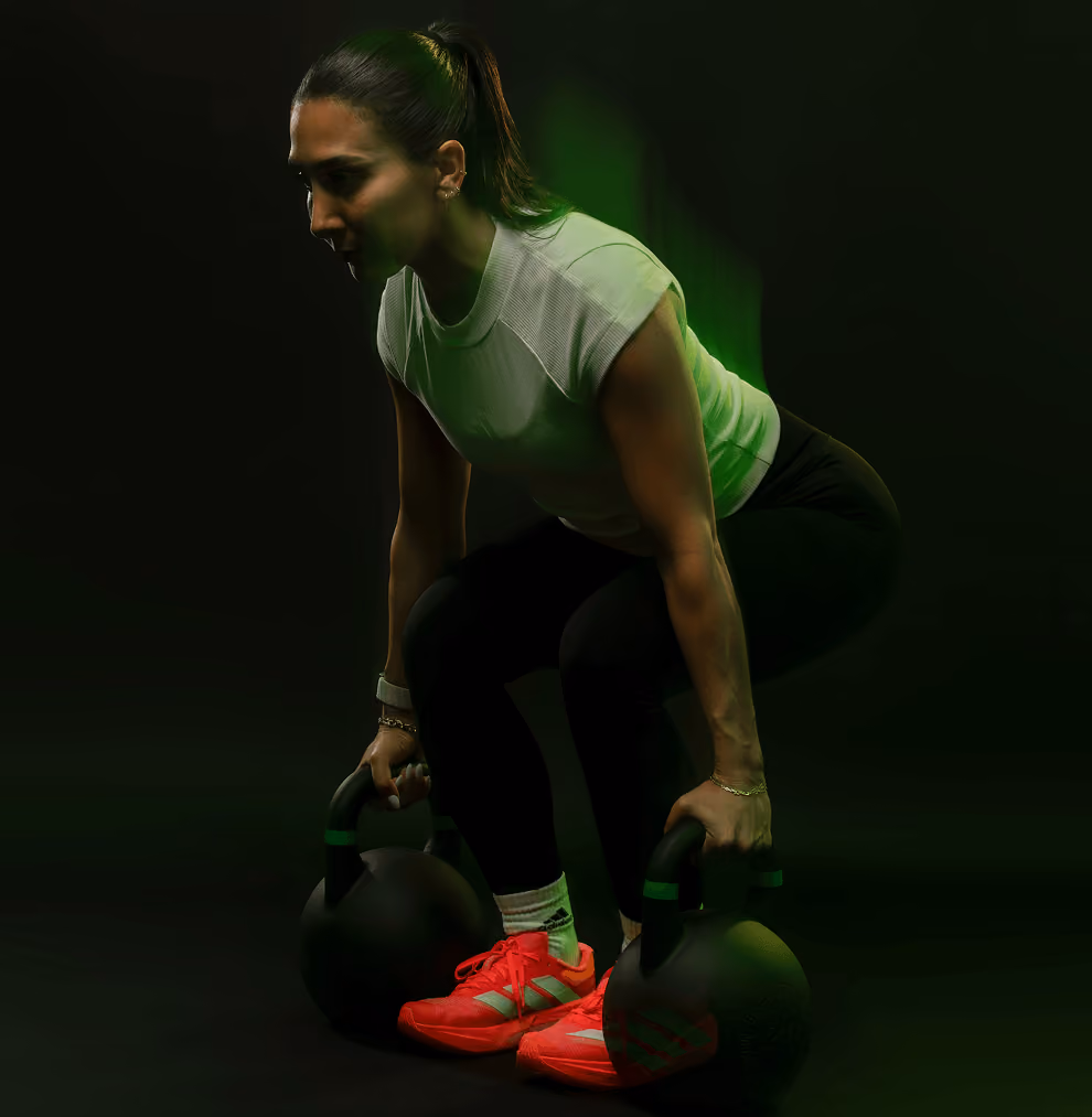 Woman in athletic attire squatting while holding two black kettlebells on a dark background, wearing bright red shoes and a white short-sleeve top.