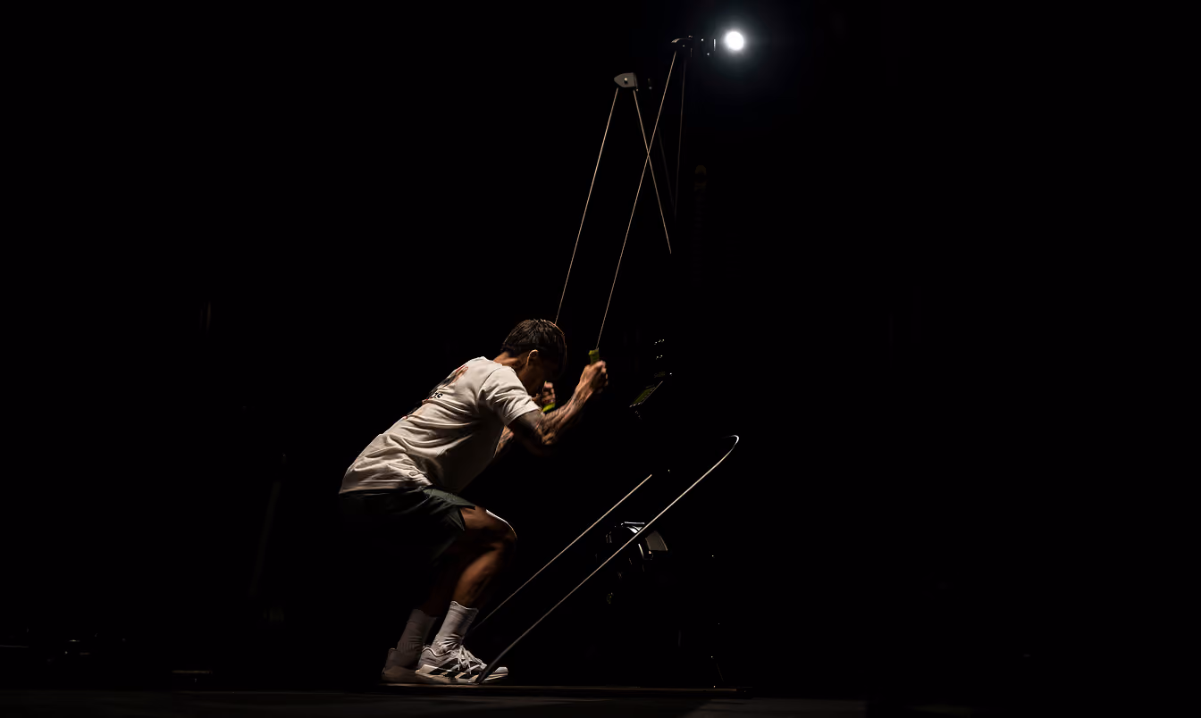 A man in a white shirt and shorts using a cable machine for resistance training in a dark gym with a bright light illuminating him from above.