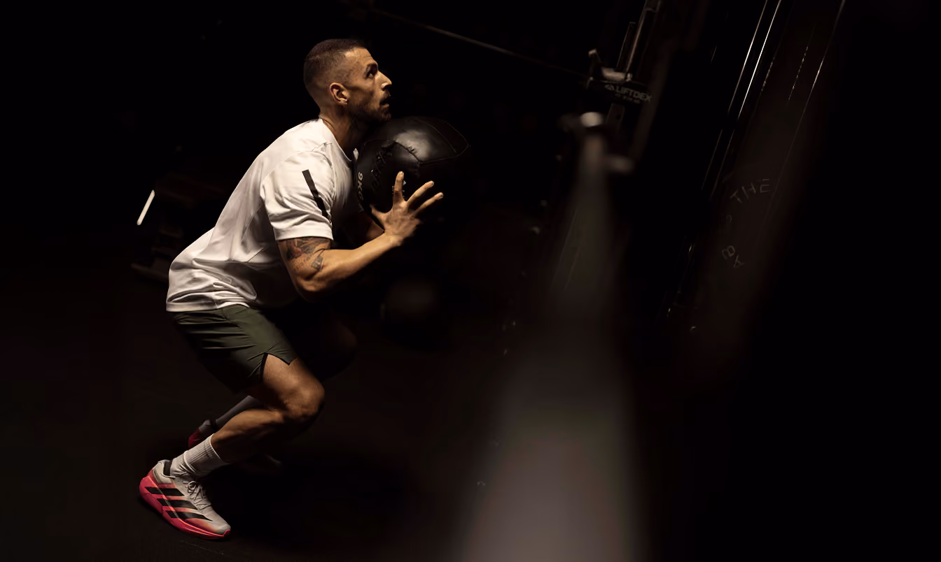 Man with short hair wearing a white shirt, green shorts, and pink and gray sneakers squats while holding a black medicine ball in a dark gym setting.