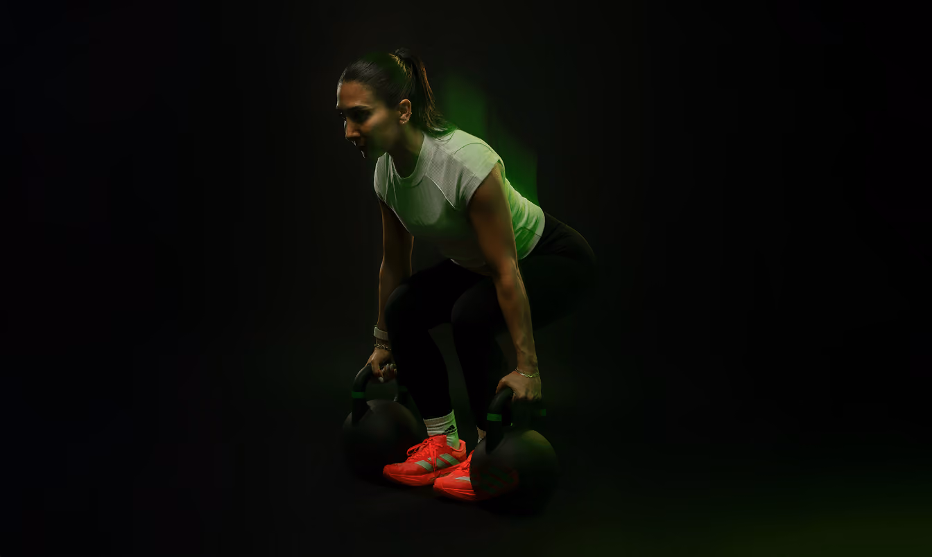 Woman in athletic attire squatting while lifting two kettlebells with a focused expression, against a dark background with green lighting accents.
