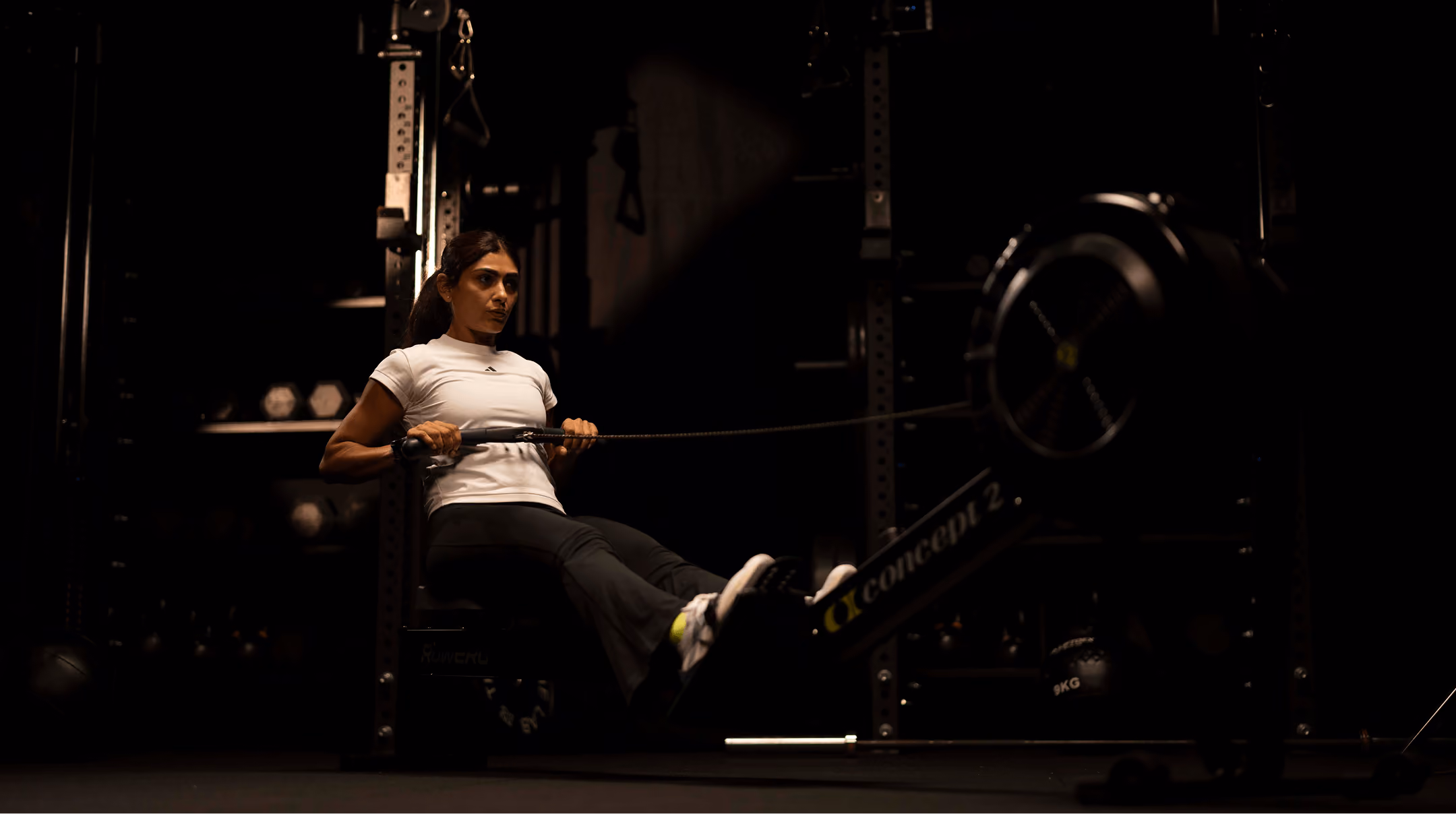 Woman in a white shirt using a rowing machine in a dark gym, focused on her workout.