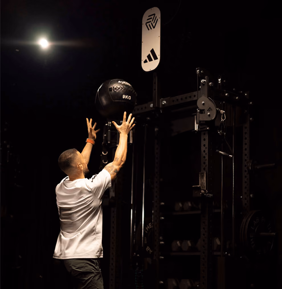 Man in white t-shirt and dark shorts throwing a 9kg black medicine ball against a padded target on a gym wall with equipment around.