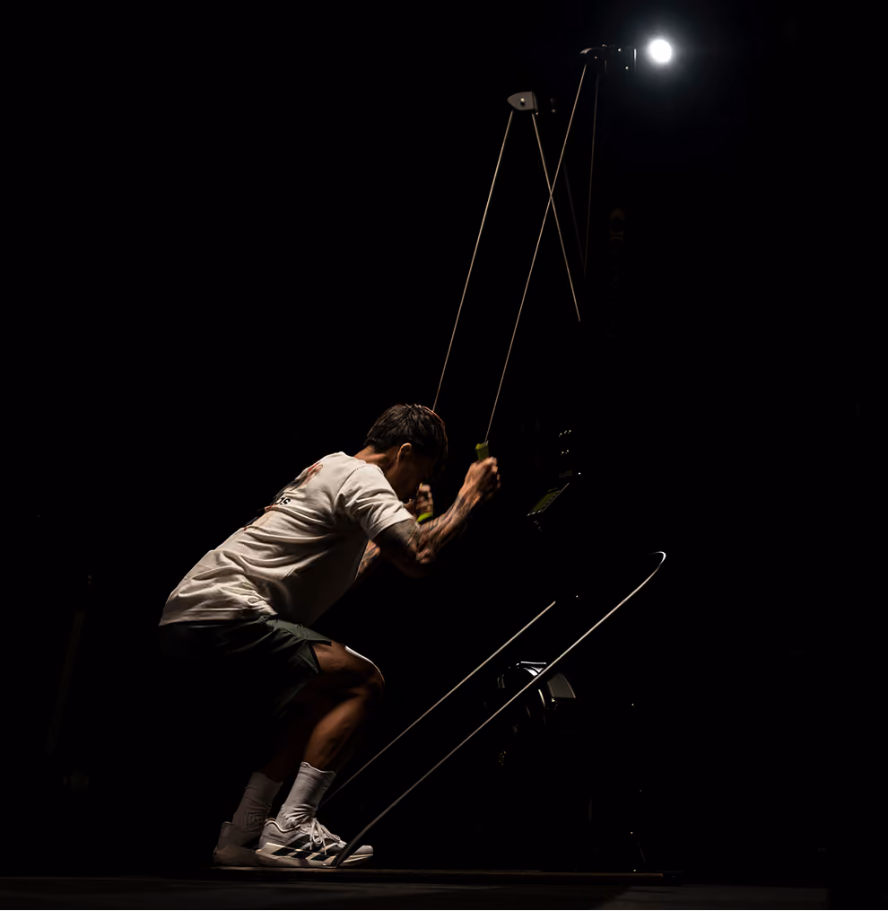 Man exercising on a ski ergometer in a dark room illuminated by a single overhead light.
