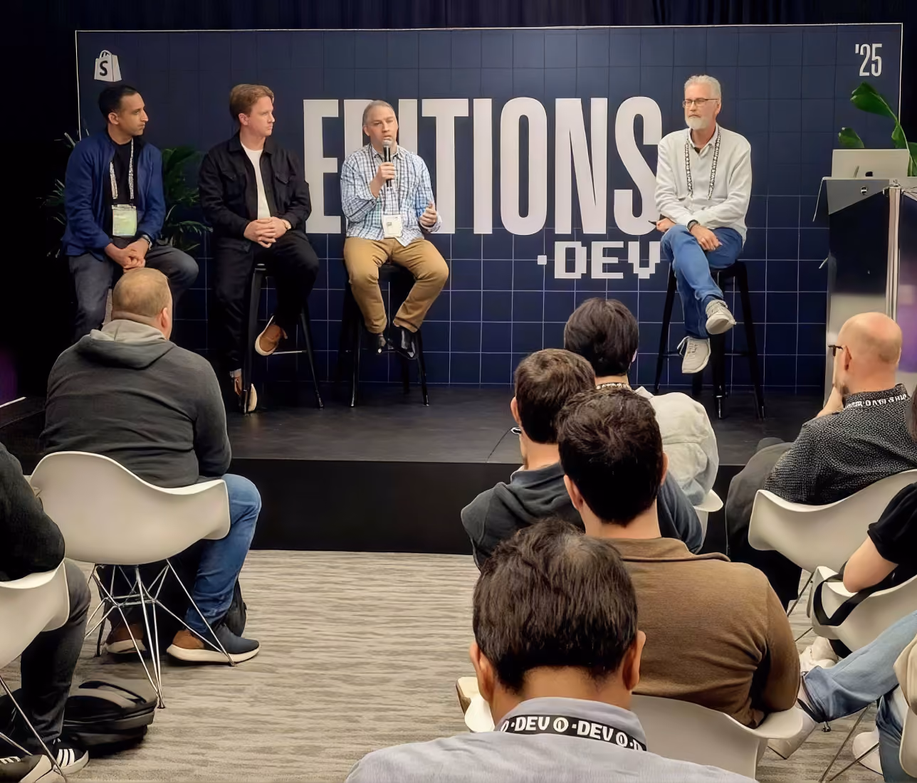 Four men seated on stools on a stage in front of an audience, one man speaking into a microphone with a backdrop showing 'EDITIONS .DEV' and '25' in the corner.