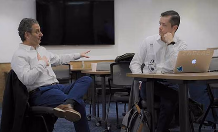 Two men in white shirts sitting and talking in an office setting with a laptop on the table and a large screen in the background.