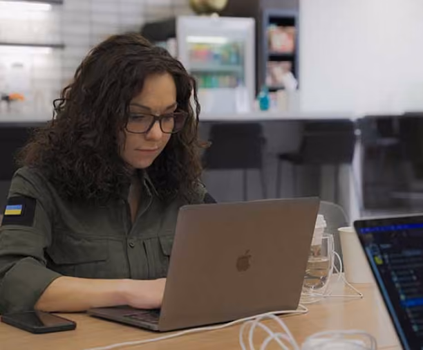 A woman with curly hair and glasses wearing a dark green shirt with a Ukrainian flag patch, working on a MacBook laptop at a table in a modern indoor setting with another laptop, phone, and cables visible.