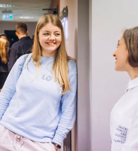 Two women smiling and conversing indoors, one in a light blue sweatshirt leaning against a wall, the other wearing a white shirt.