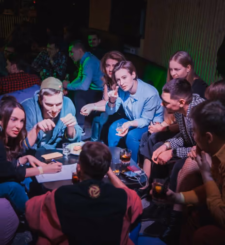 Group of young adults sitting closely around a low table in a dimly lit room, engaged in conversation and holding drinks, some writing on paper, with casual expressions.