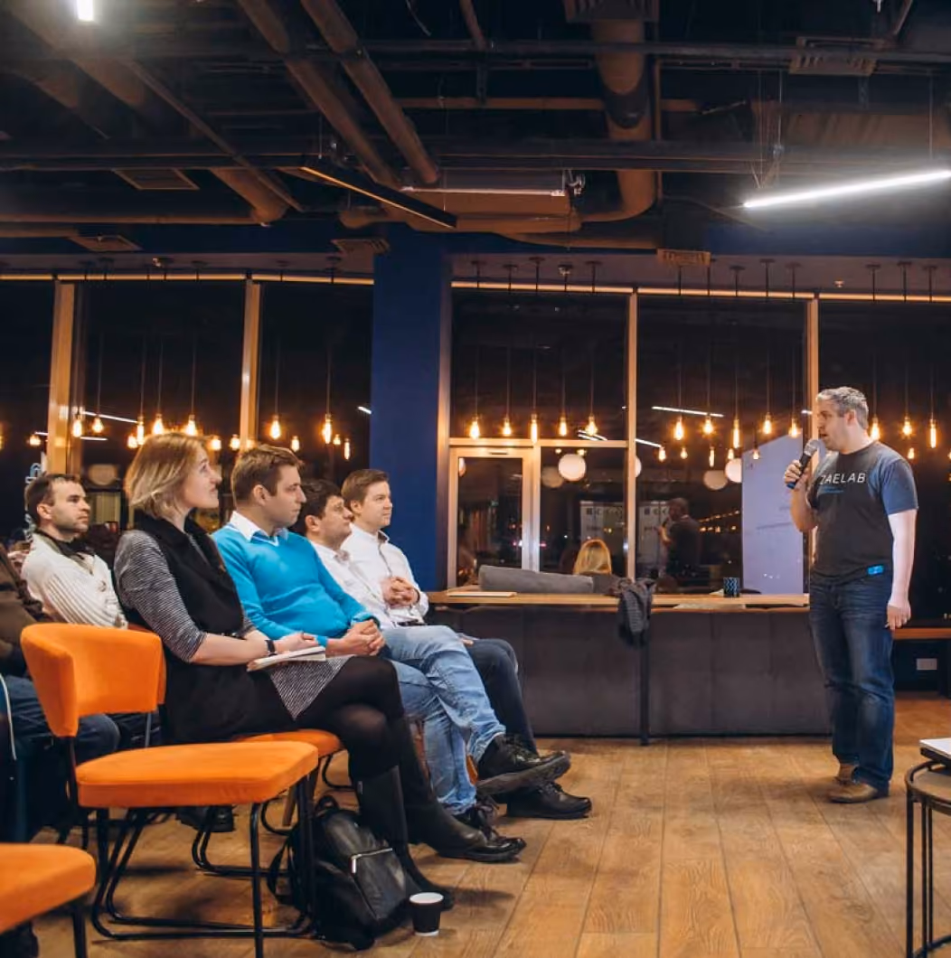 A man holding a microphone is speaking to an audience seated in orange chairs in a modern room with wooden floors and hanging lights.
