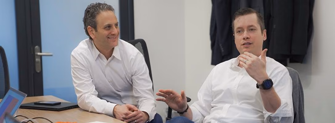 Two men wearing white shirts engaged in conversation while seated at a conference table in an office setting.