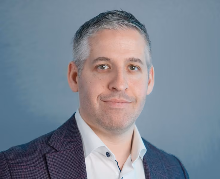 Portrait of a middle-aged man with short gray hair and light skin wearing a white shirt and a dark checkered blazer, smiling slightly against a plain gray background.